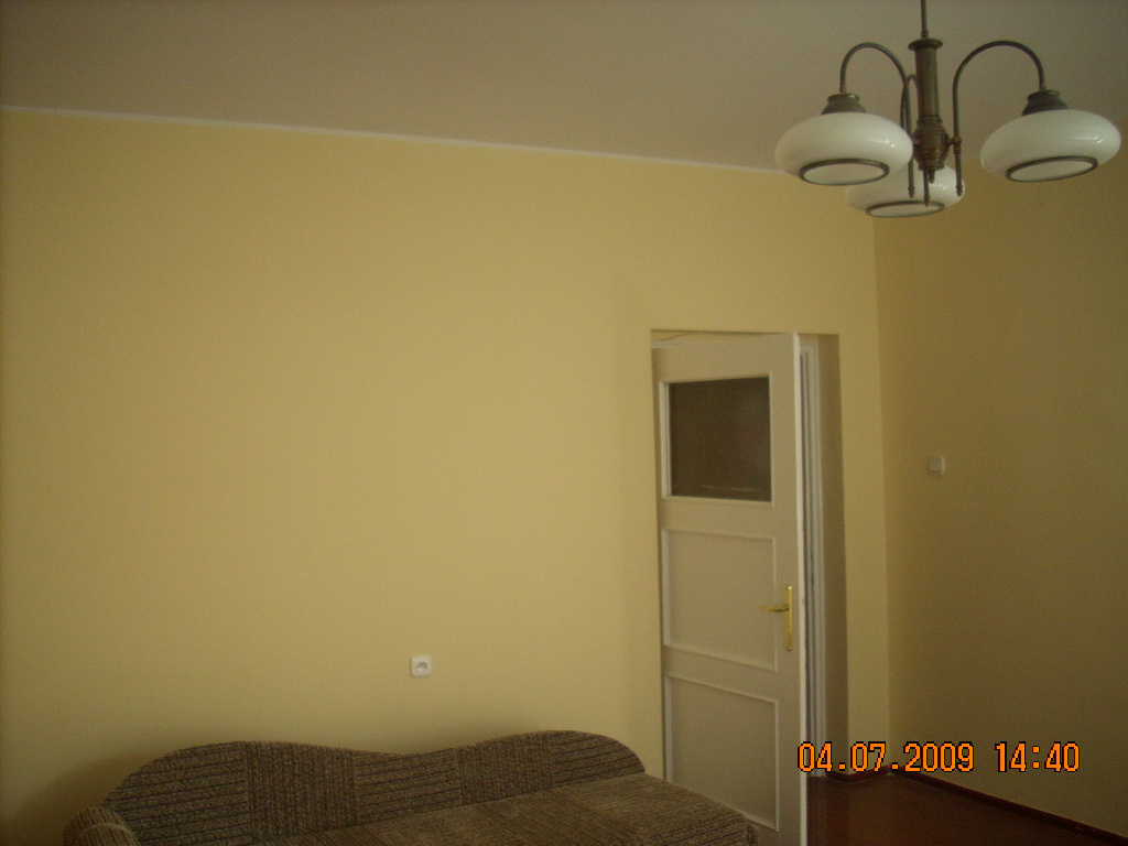 Żółta ściana w pokoju z beżową kanapą, widoczna biała futryna otwartych drzwi, lampa sufitowa z trzema białymi kloszami i gniazdko elektryczne na ścianie.