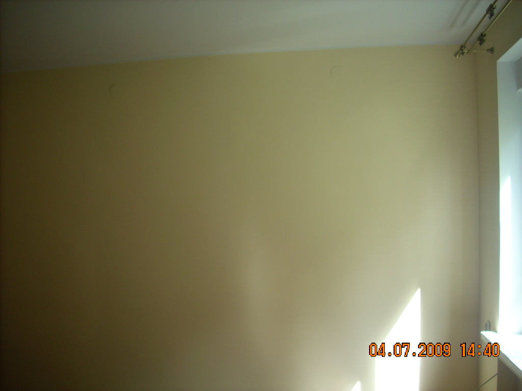 Ściana pomalowana na jasnożółty kolor z widocznymi śladami po wcześniejszych mocowaniach, widoczne na górze ściany w Poznaniu, z fragmentem okna po prawej stronie.