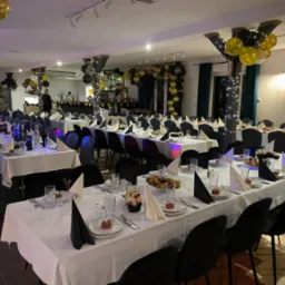 Uroczysta sala z długimi stołami nakrytymi białymi obrusami, czarnymi krzesłami i dekoracjami z czarno-złotych balonów, przygotowana na przyjęcie.
