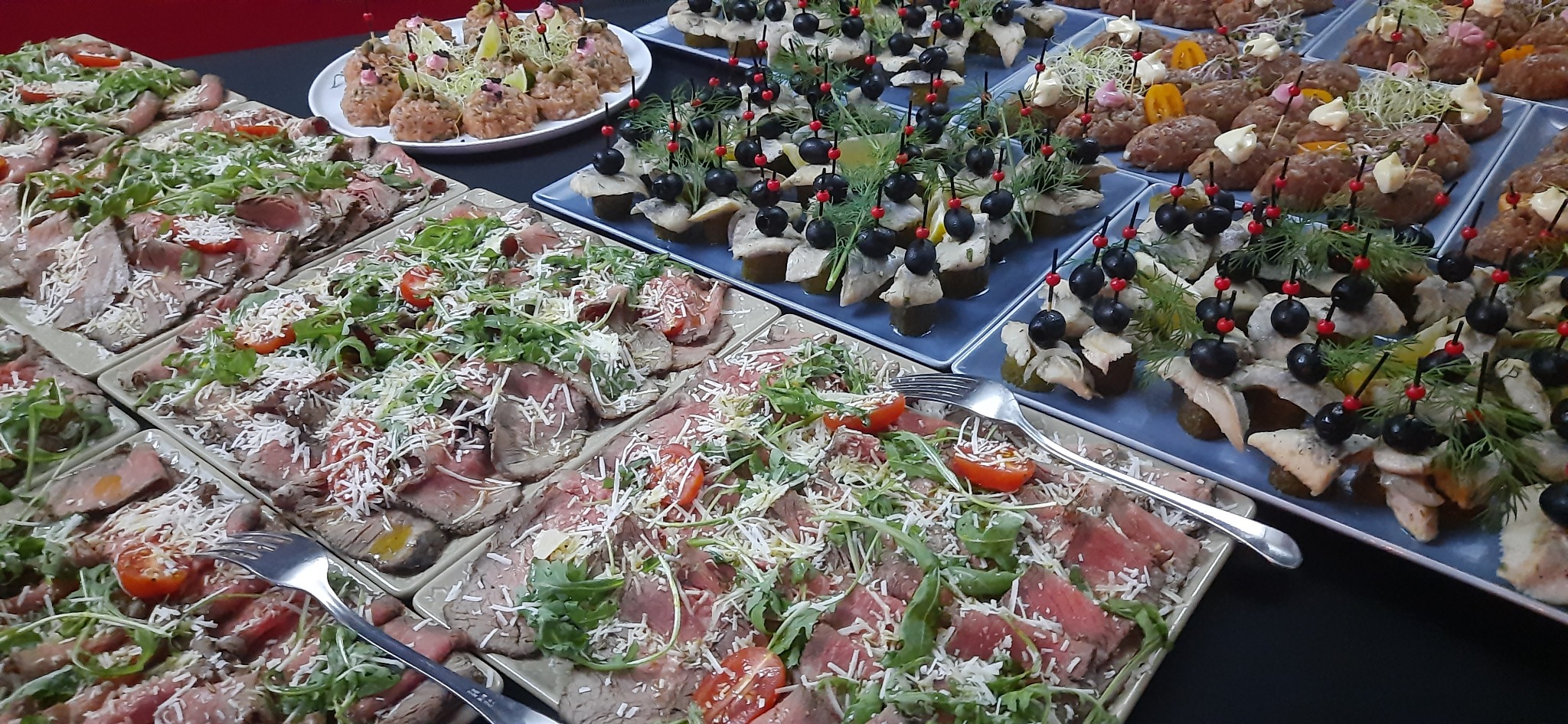 Rozmaite przekąski na paterach: plastry pieczonego mięsa z rukolą i pomidorami, koreczki ze śledzia z oliwkami i ogórkiem, oraz kulki mięsne z dekoracją, gotowe do serwowania.