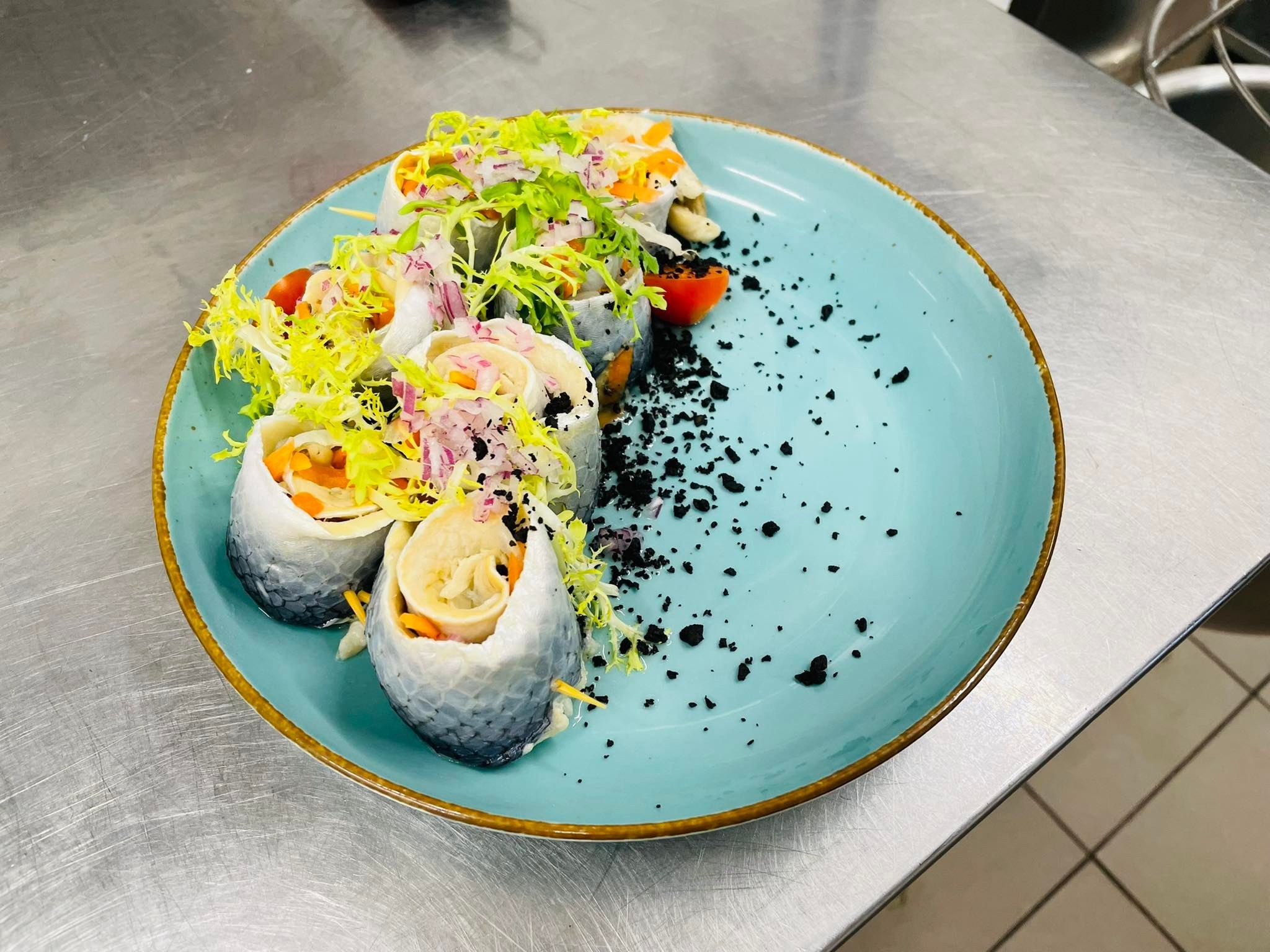 Rolmopsy z dodatkiem warzyw i ziół, podane na błękitnym talerzu z brązową obwódką, posypane czarnym proszkiem, widoczne z bliska na metalowym stole.