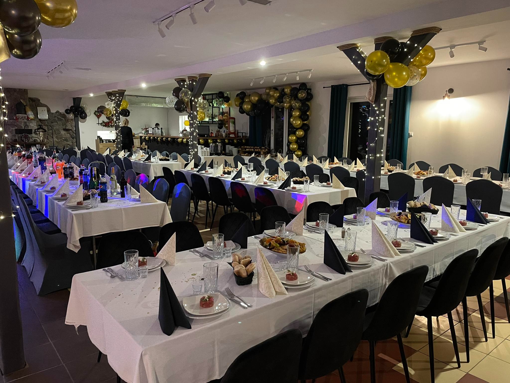 Uroczysta sala z długimi stołami nakrytymi białymi obrusami, czarnymi krzesłami i dekoracjami z czarno-złotych balonów, przygotowana na przyjęcie.