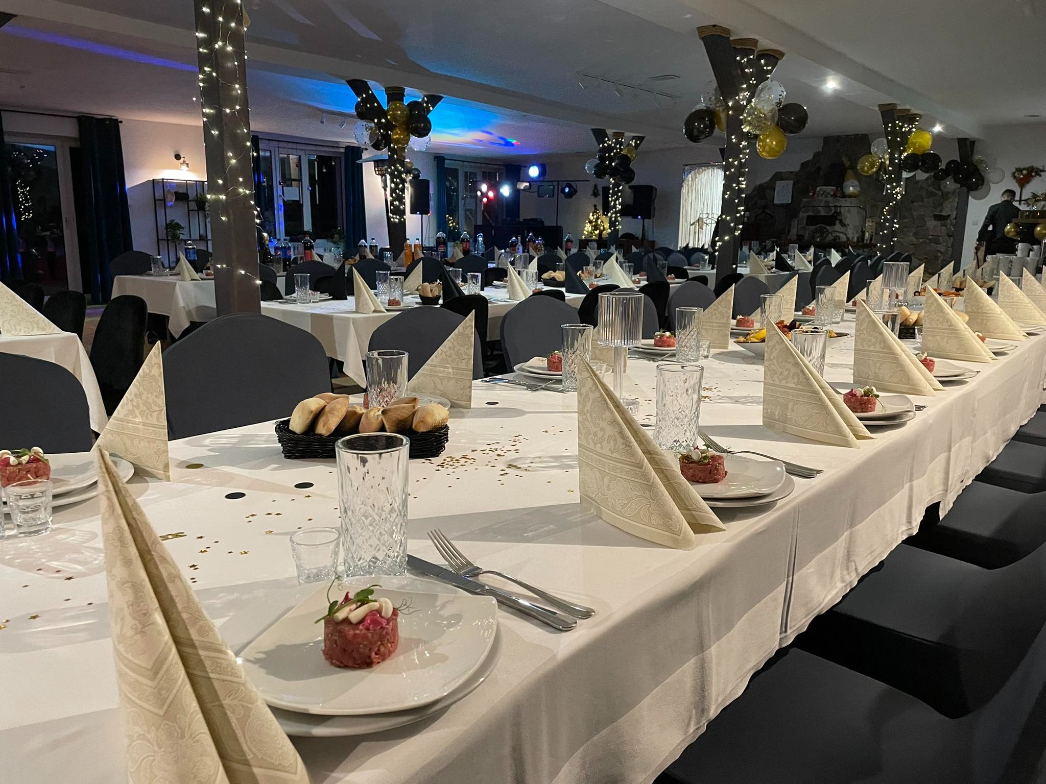 Uroczysta sala bankietowa z długimi stołami nakrytymi białymi obrusami, elegancko złożonymi serwetkami, szklankami z wzorem i przystawkami na talerzach, dekoracje z balonów i światełek na filarach.
