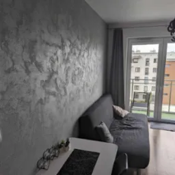 Ściana w salonie wykończona dekoracyjną, srebrzystą gładzią w stylu weneckim, widoczny fragment sofy, stolika i balkonu.