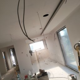 Osoba prywatna Bartłomiej Mainka - Wnętrze w trakcie wykańczania: widoczne przewody elektryczne zwisające z sufitu, gładkie, białe ściany, szlifowanie ściany przy oknie, drabina oraz materiały budowlane na podłodze.
