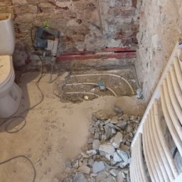 Osoba prywatna Bartłomiej Mainka - Remont łazienki: kucie podłoża pod instalację ogrzewania podłogowego. Widoczne rury, gruz i młot udarowy przy starej ceglanej ścianie.