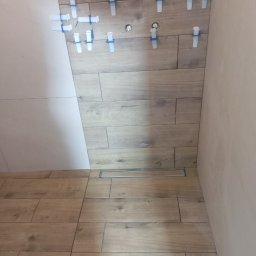 Osoba prywatna Bartłomiej Mainka - Układanie płytek imitujących drewno na ścianie i podłodze w łazience. Widoczne klipsy poziomujące i odpływ liniowy. Część ściany wykończona białą płytką.