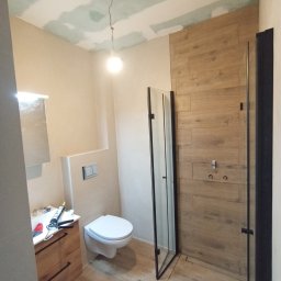 Osoba prywatna Bartłomiej Mainka - Remont łazienki: Ukończona kabina prysznicowa z drewnopodobnymi płytkami, toaleta podwieszana i umywalka z drewnianą szafką. Sufit z płyt karton-gips.