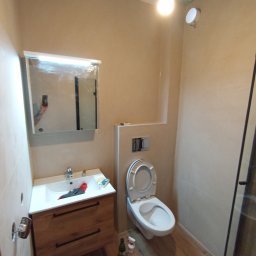 Osoba prywatna Bartłomiej Mainka - Wykończona łazienka z drewnianą szafką pod umywalką, toaletą i lustrem. Widoczne narzędzia i butelki wskazują na trwające prace wykończeniowe.