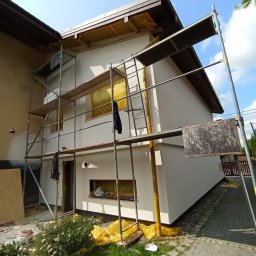 Osoba prywatna Bartłomiej Mainka - Elewacja domu w trakcie malowania, widoczne rusztowania, zabezpieczone okna folią. Świeżo pomalowana ściana w kolorze białym. Słoneczny dzień.