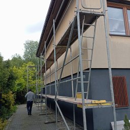 Bart-Dom - Dom w trakcie prac elewacyjnych z użyciem rusztowania. Widoczny fragment ściany w dwóch odcieniach, okno i osoba oddalająca się wzdłuż budynku.