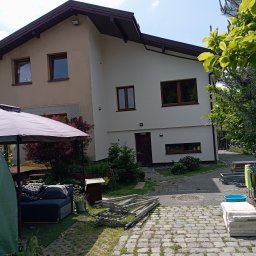 Osoba prywatna Bartłomiej Mainka - Dwukondygnacyjny dom z beżową i białą elewacją, brązowymi oknami, widoczny fragment ogrodu z roślinnością i rozłożone rusztowanie. Słoneczny dzień.