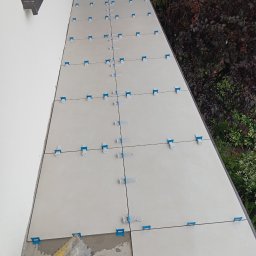 Osoba prywatna Bartłomiej Mainka - Układanie jasnych płytek na zewnątrz budynku z użyciem systemu poziomowania, widoczne narzędzia glazurnicze i zieleń w tle. Prace w toku.