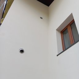 Osoba prywatna Bartłomiej Mainka - Jasna ściana po malowaniu z kratką wentylacyjną i oknem z brązową ramą. Widoczne rusztowanie z folią ochronną. Ciemny sufit w rogu.