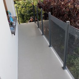 Osoba prywatna Bartłomiej Mainka - Nowoczesny balkon z szarą szklaną balustradą i jasnymi płytkami, widok na zieleń. Dwóch mężczyzn montuje balustradę.