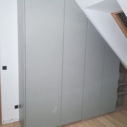 Bart-Dom - Nowoczesna zabudowa szafy pod skosem dachu, z matowymi, jasnoszarymi frontami bez uchwytów, harmonijnie wkomponowana w przestrzeń poddasza z drewnianą podłogą.