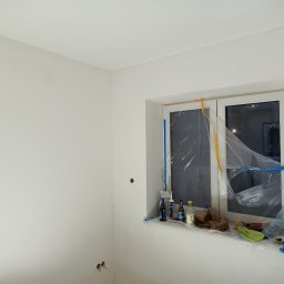 Bart-Dom - Gładkie, białe ściany w pomieszczeniu podczas prac wykończeniowych. Okno zabezpieczone folią i niebieską taśmą, na parapecie butelki, tuby z uszczelniaczem, pomarańczowa taśma i zielona szmatka.