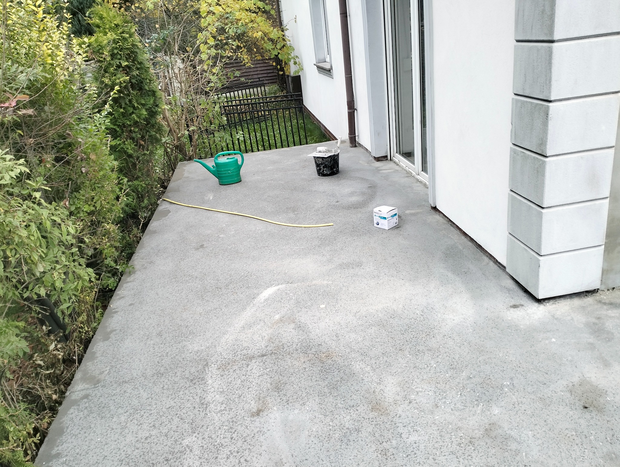 Przygotowany do malowania betonowy taras z widocznymi akcesoriami: konewka, wiadro z farbą i puszka farby. Tło stanowi biała ściana budynku i zieleń.