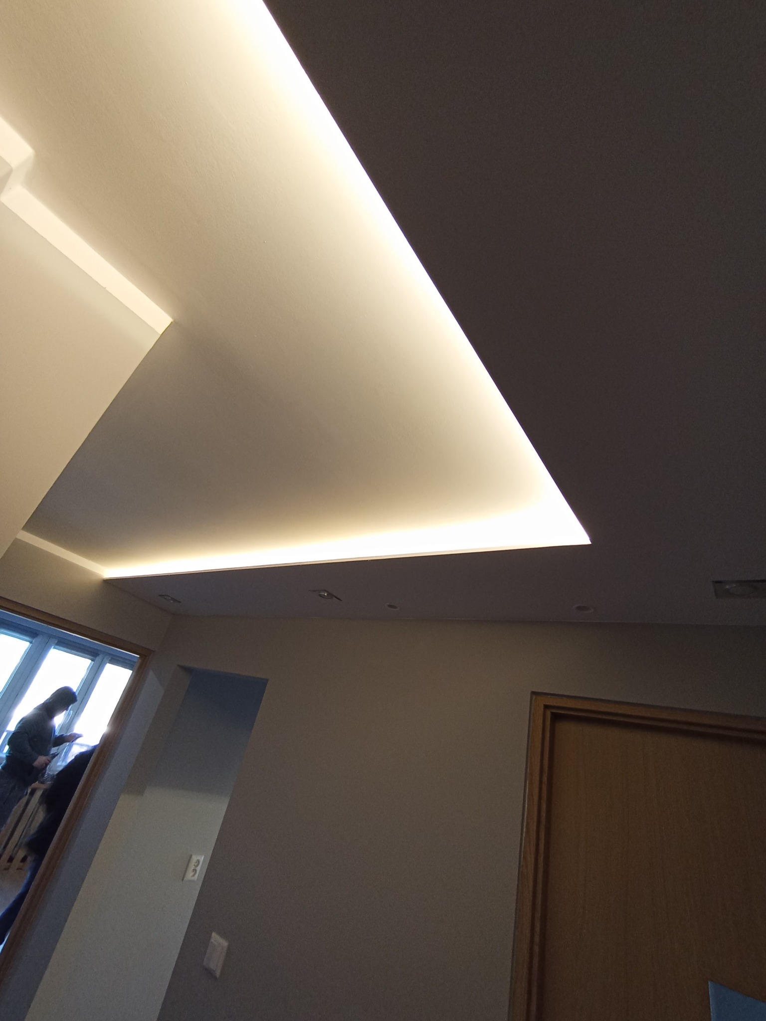 Nowoczesny sufit podwieszany z oświetleniem LED w jasnym wnętrzu. Widok na okno i fragment drzwi. Minimalistyczny design, stonowana kolorystyka.