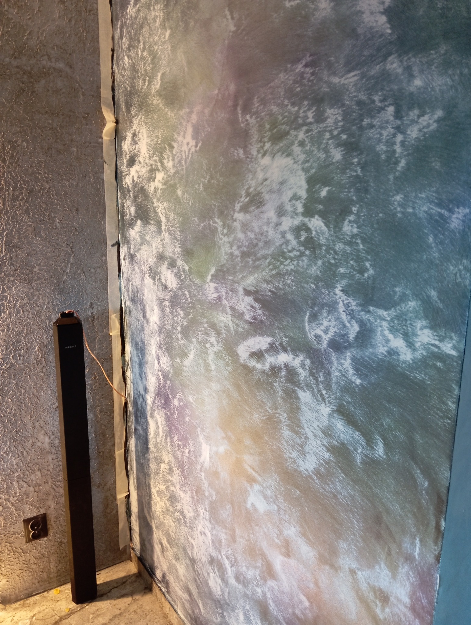 Ściana z dekoracyjną, wielobarwną fakturą, zabezpieczona taśmą malarską przy krawędziach. Obok stoi czarny głośnik, widoczne gniazdko elektryczne.