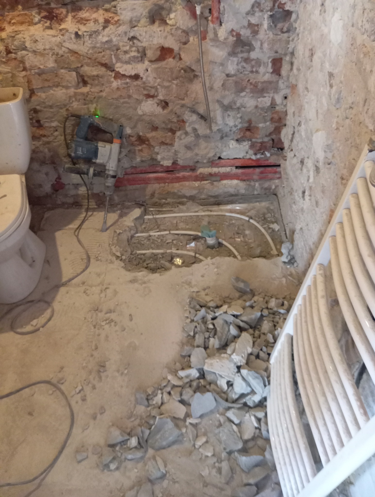 Remont łazienki: kucie podłoża pod instalację ogrzewania podłogowego. Widoczne rury, gruz i młot udarowy przy starej ceglanej ścianie.