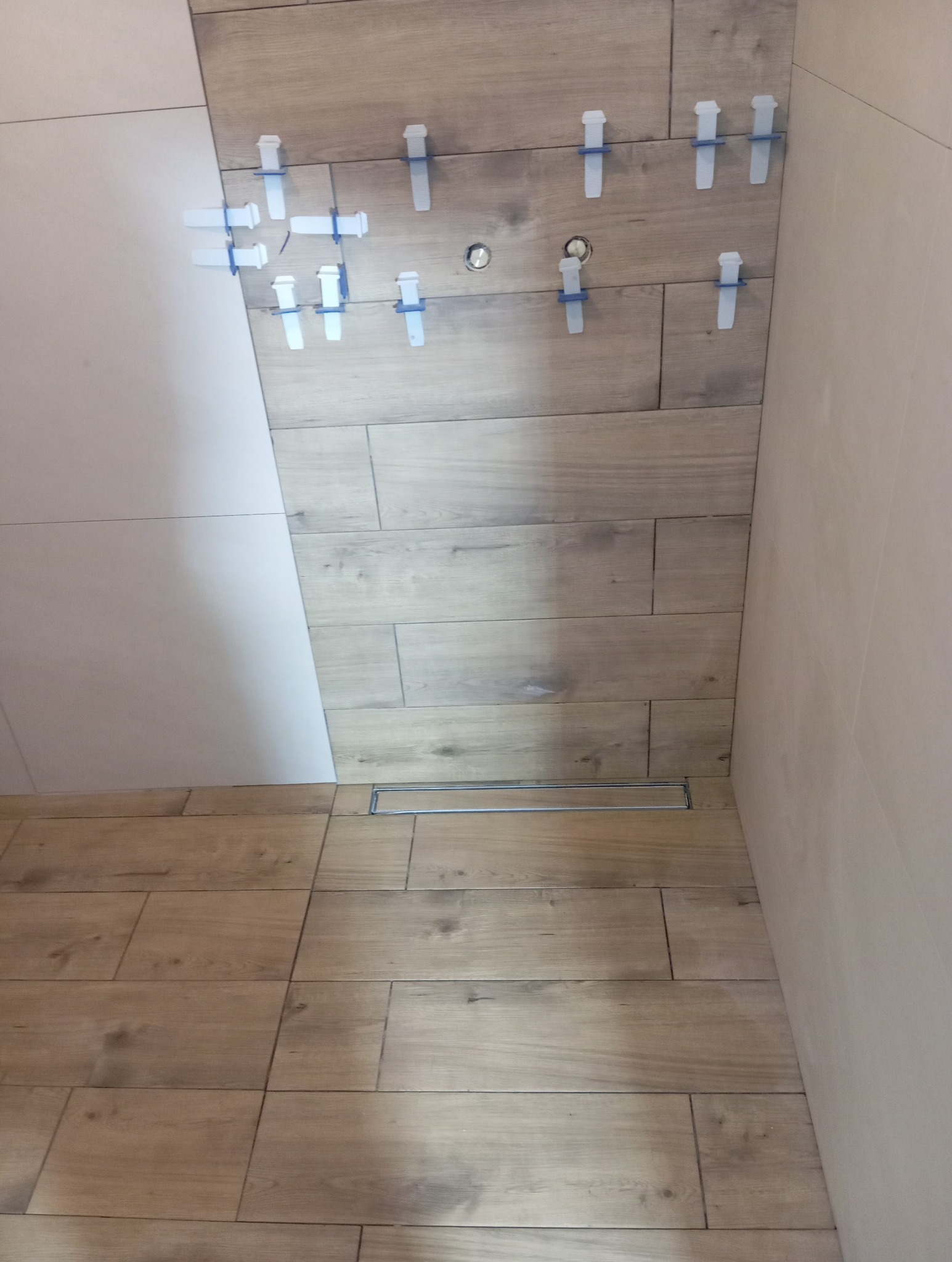 Układanie płytek imitujących drewno na ścianie i podłodze w łazience. Widoczne klipsy poziomujące i odpływ liniowy. Część ściany wykończona białą płytką.