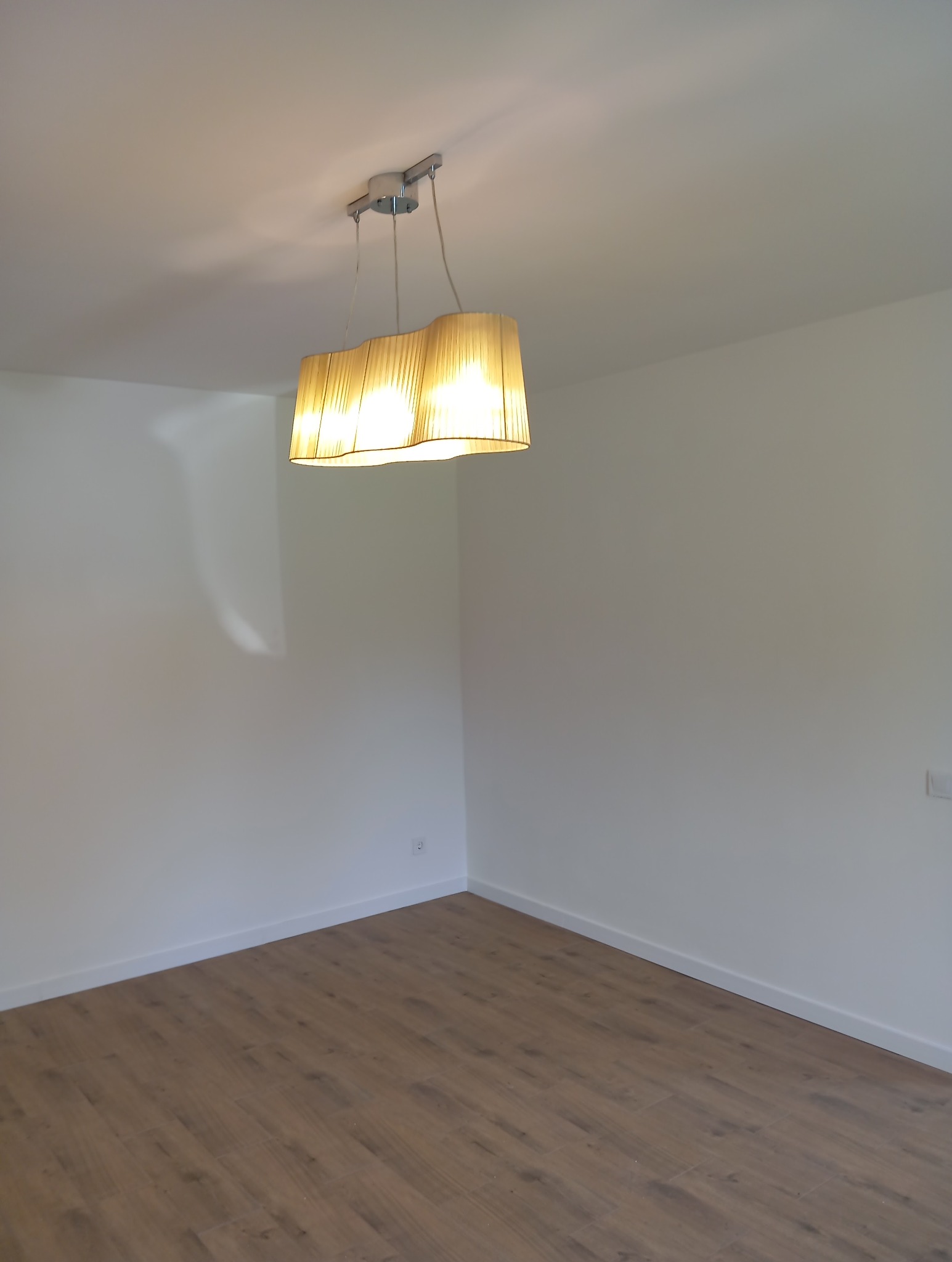 Jasne panele podłogowe w nowym wnętrzu z białymi ścianami i wiszącą lampą z abażurem. Widoczne gniazdko elektryczne.
