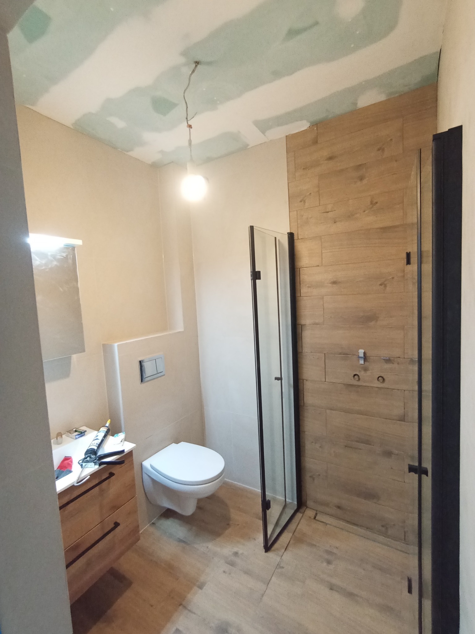 Remont łazienki: Ukończona kabina prysznicowa z drewnopodobnymi płytkami, toaleta podwieszana i umywalka z drewnianą szafką. Sufit z płyt karton-gips.