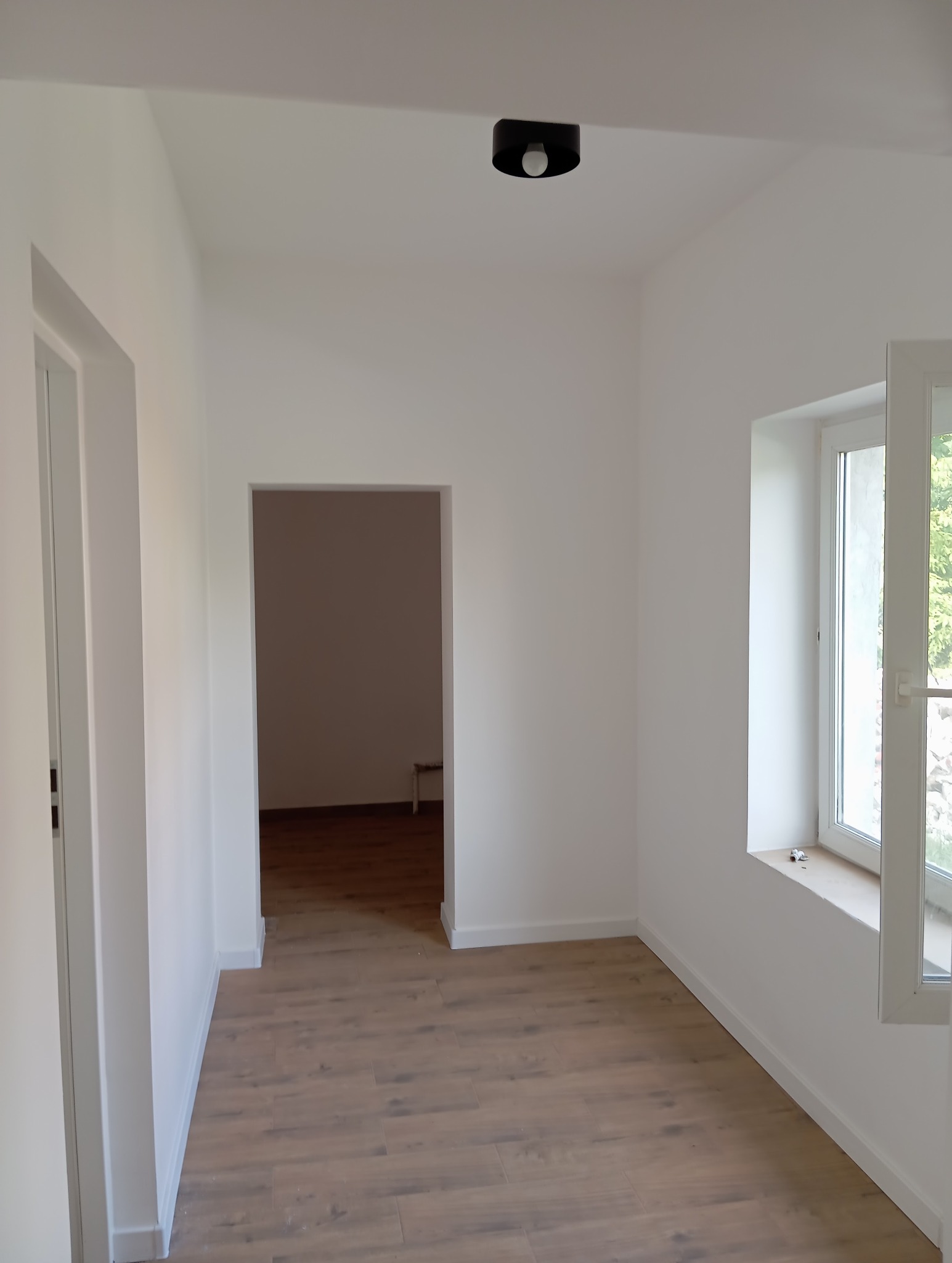 Wnętrze po remoncie: białe ściany, drewnopodobna podłoga, okno z widokiem na zieleń. Minimalistyczna lampa sufitowa, otwarte drzwi do kolejnego pomieszczenia.