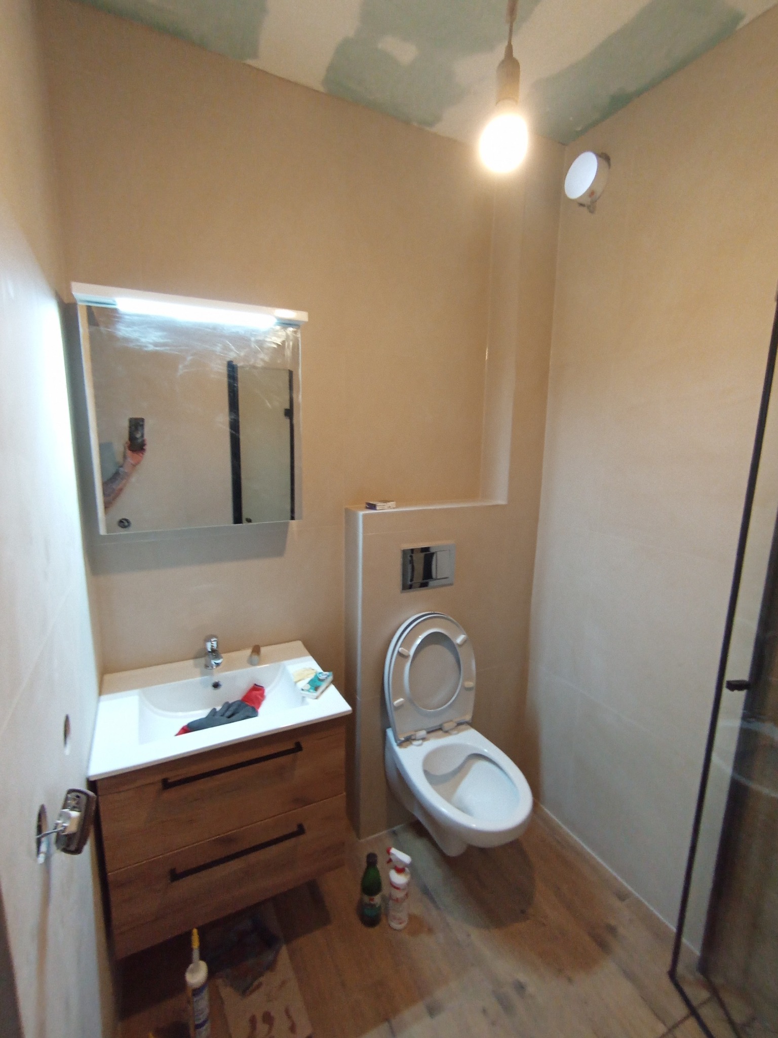 Wykończona łazienka z drewnianą szafką pod umywalką, toaletą i lustrem. Widoczne narzędzia i butelki wskazują na trwające prace wykończeniowe.