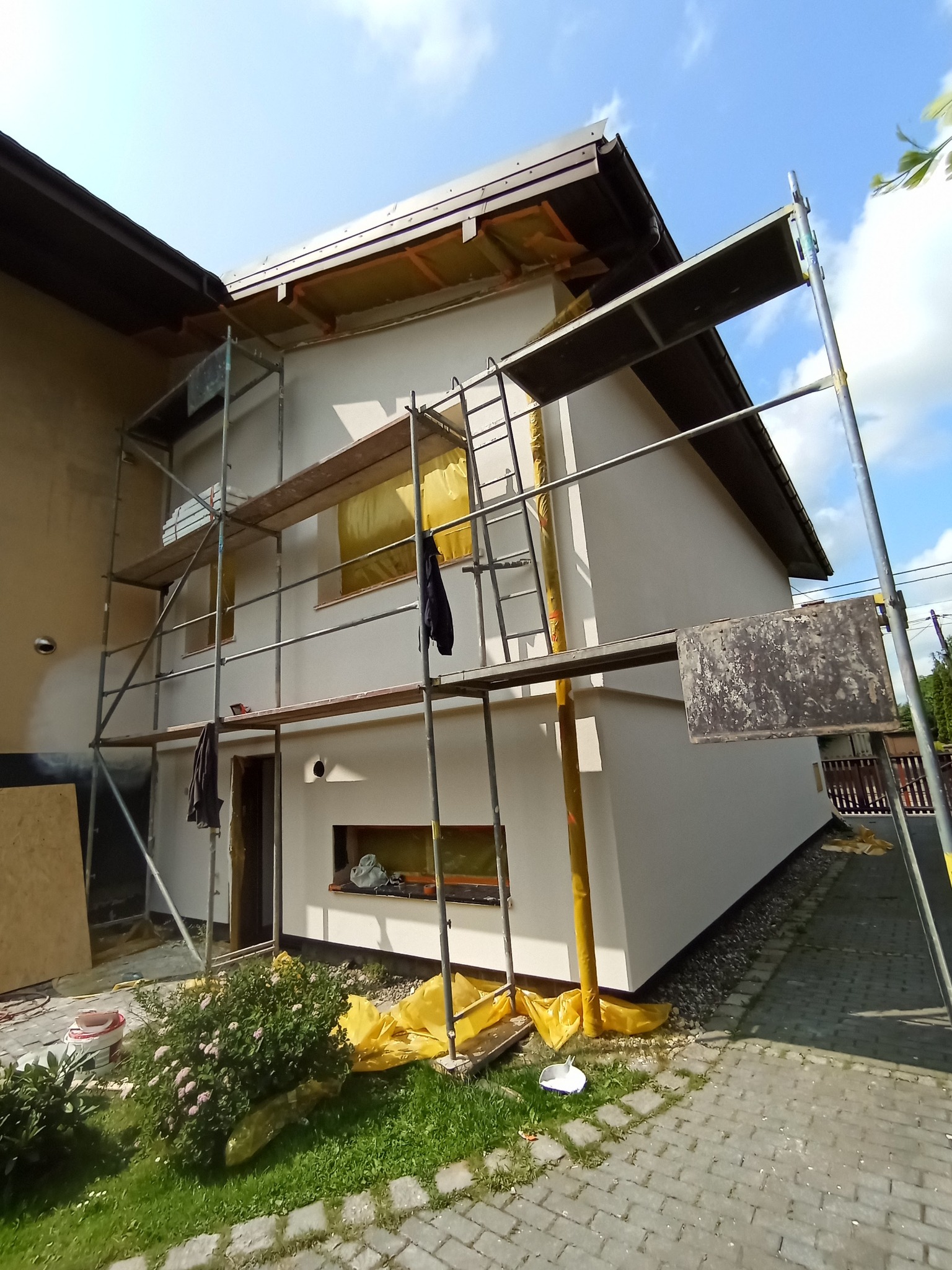 Elewacja domu w trakcie malowania, widoczne rusztowania, zabezpieczone okna folią. Świeżo pomalowana ściana w kolorze białym. Słoneczny dzień.