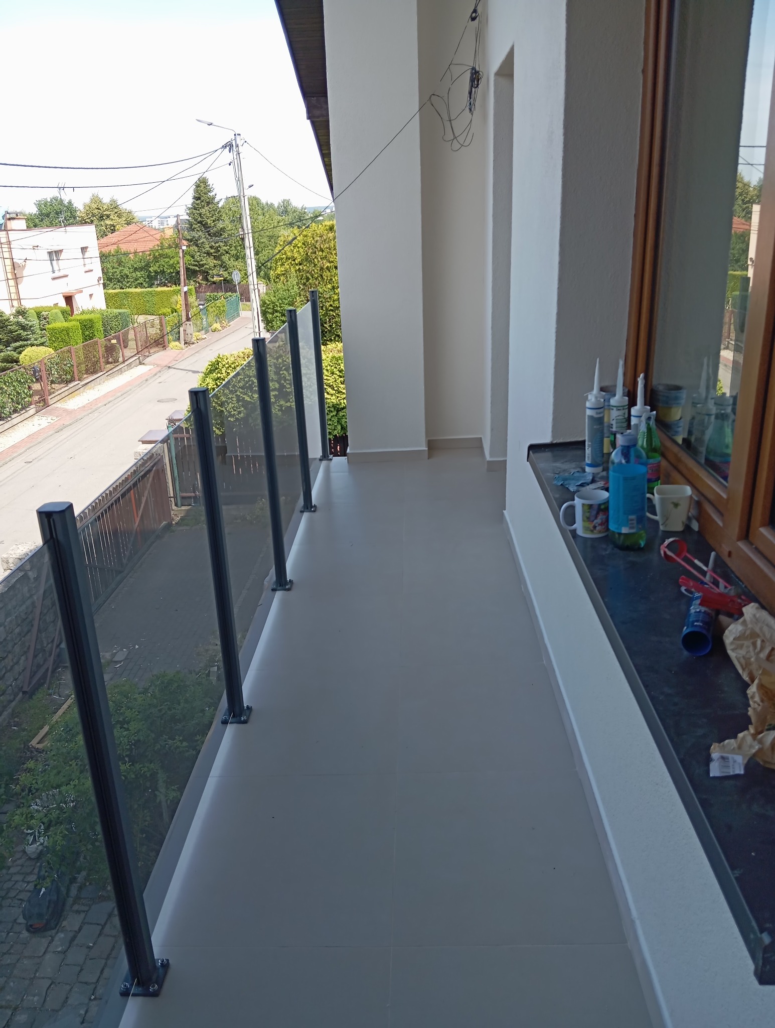 Wykończony balkon z szklaną balustradą i jasnymi płytkami. Na parapecie widoczne narzędzia i materiały budowlane. Widok na ulicę z zielenią.