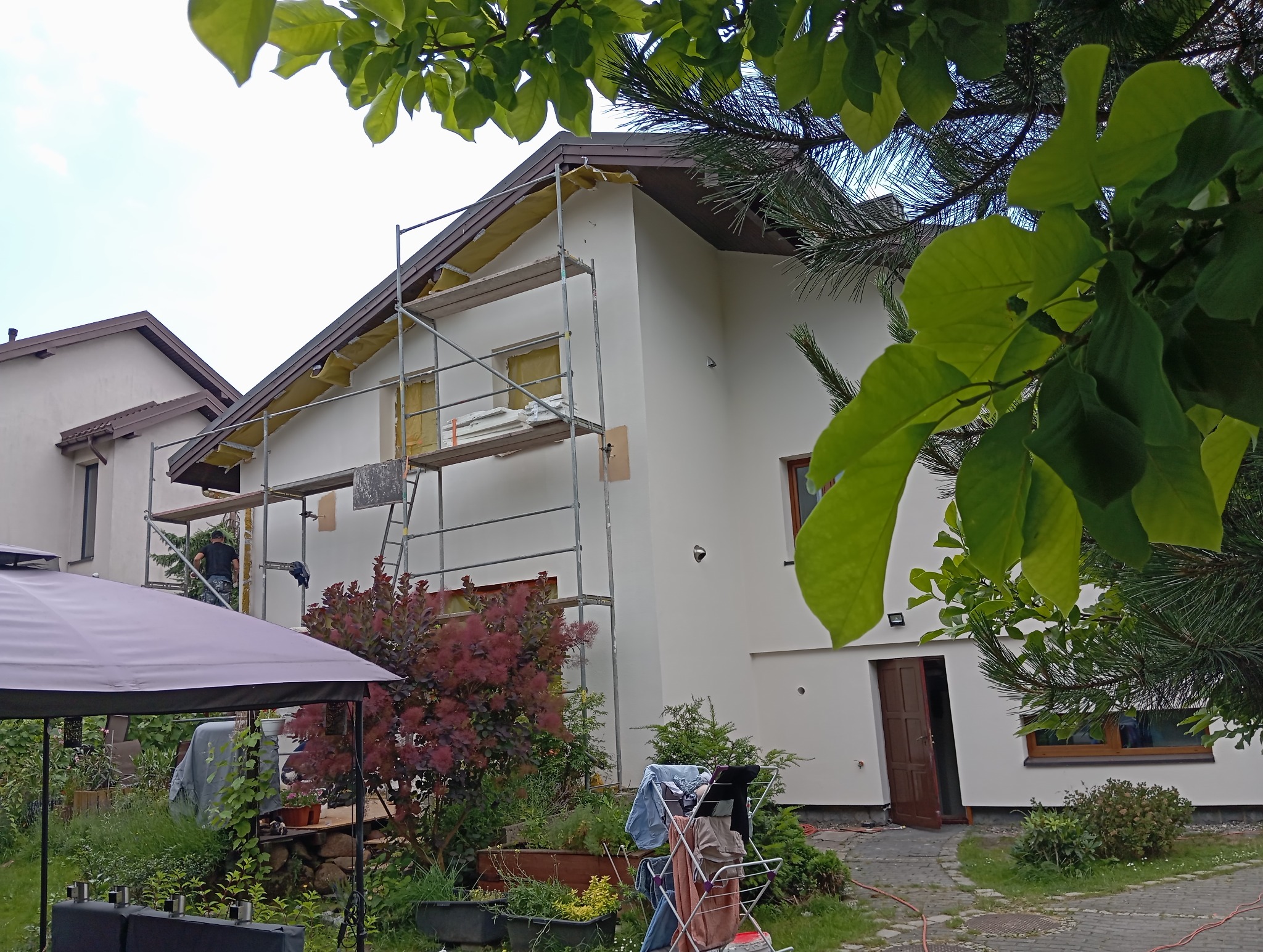 Ocieplanie i malowanie elewacji domu jednorodzinnego. Na rusztowaniu pracuje osoba, widoczne materiały izolacyjne i narzędzia. W tle zieleń ogrodu.