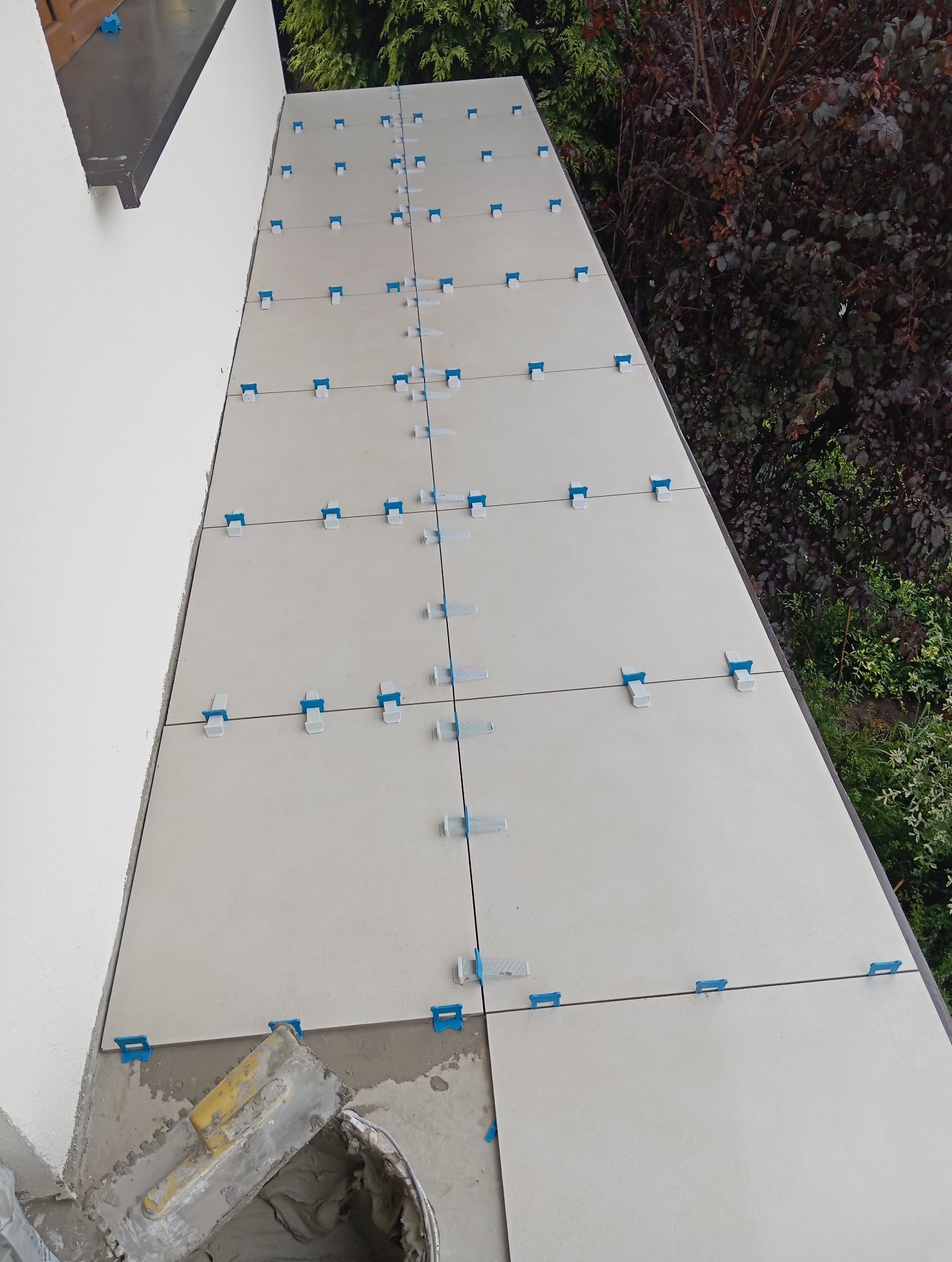 Układanie jasnych płytek na zewnątrz budynku z użyciem systemu poziomowania, widoczne narzędzia glazurnicze i zieleń w tle. Prace w toku.