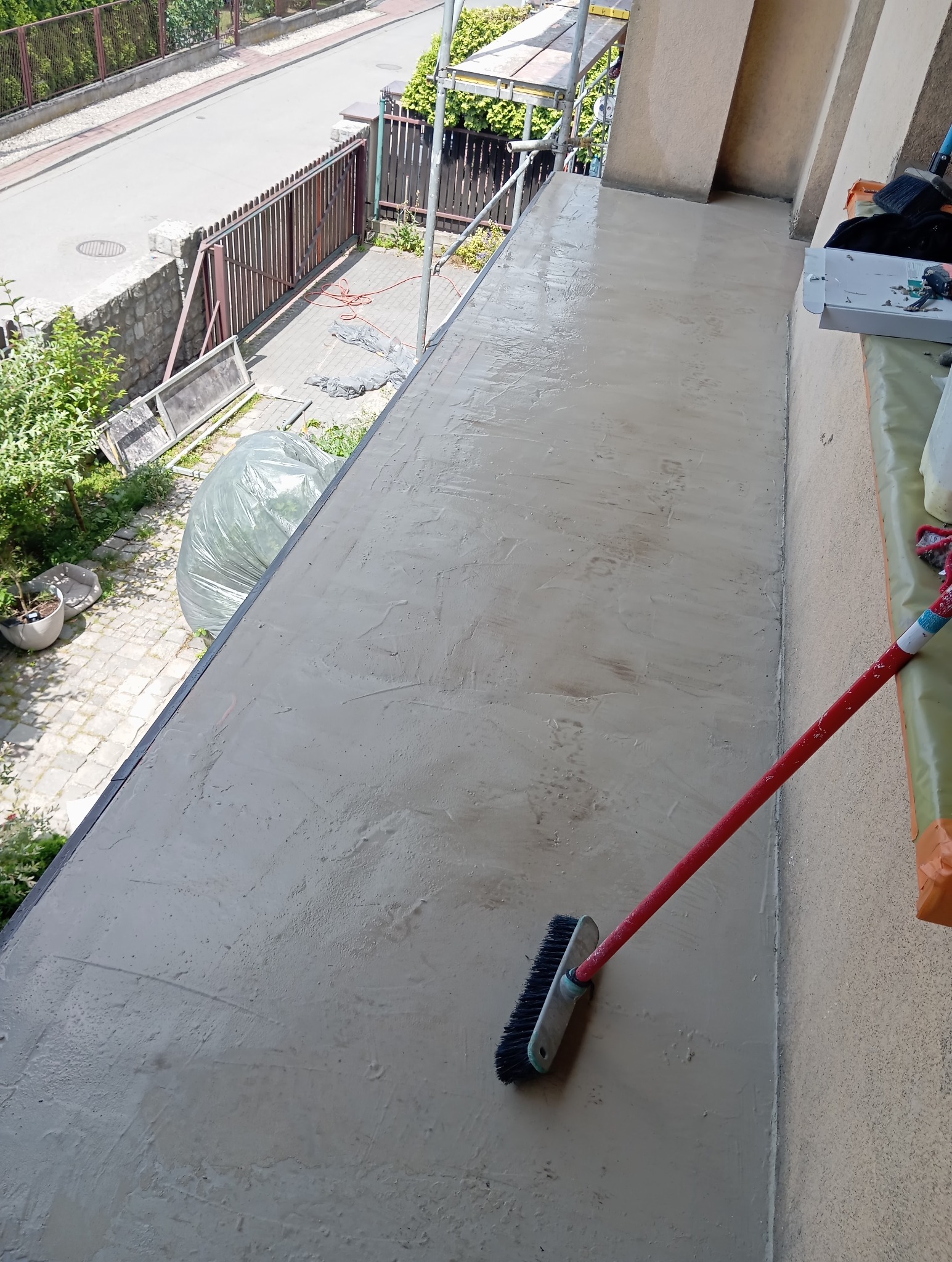 Balkon po wylewce wyrównującej, widoczna szczotka na czerwonym trzonku. W tle rusztowanie i fragment ulicy.