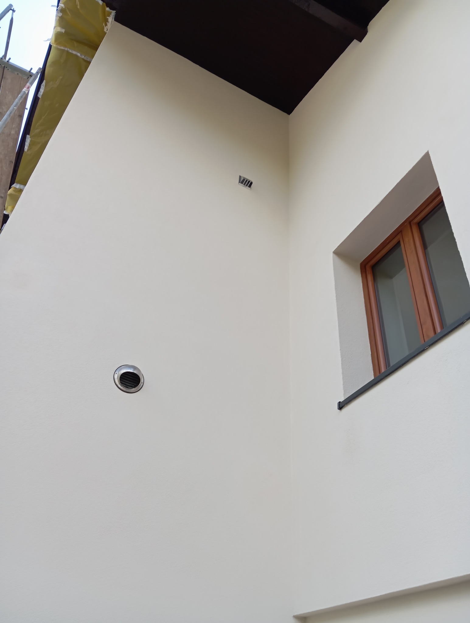 Jasna ściana po malowaniu z kratką wentylacyjną i oknem z brązową ramą. Widoczne rusztowanie z folią ochronną. Ciemny sufit w rogu.