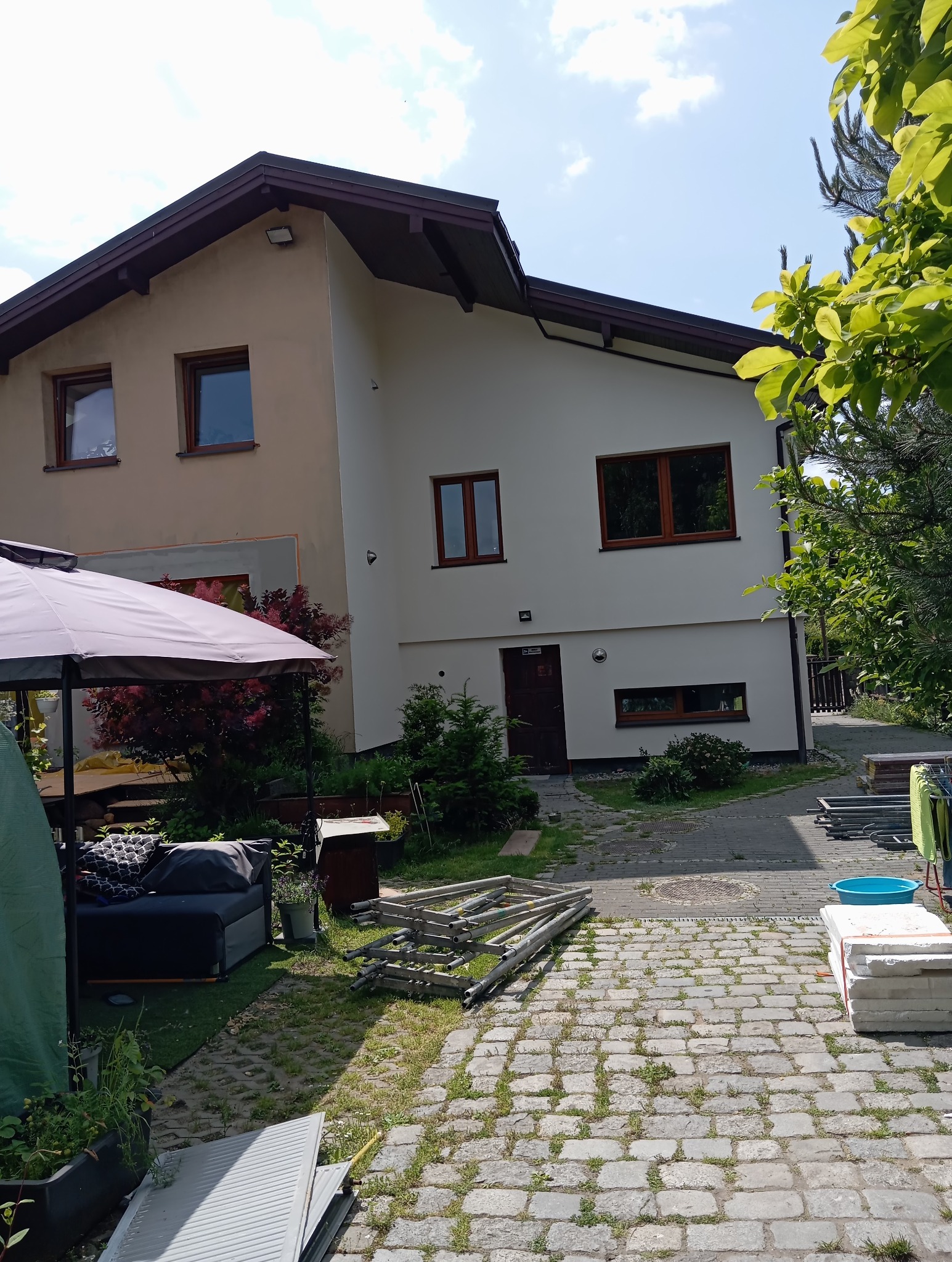 Dwukondygnacyjny dom z beżową i białą elewacją, brązowymi oknami i dachem. Na pierwszym planie widoczne rusztowania i materiały budowlane na brukowanym podwórku.