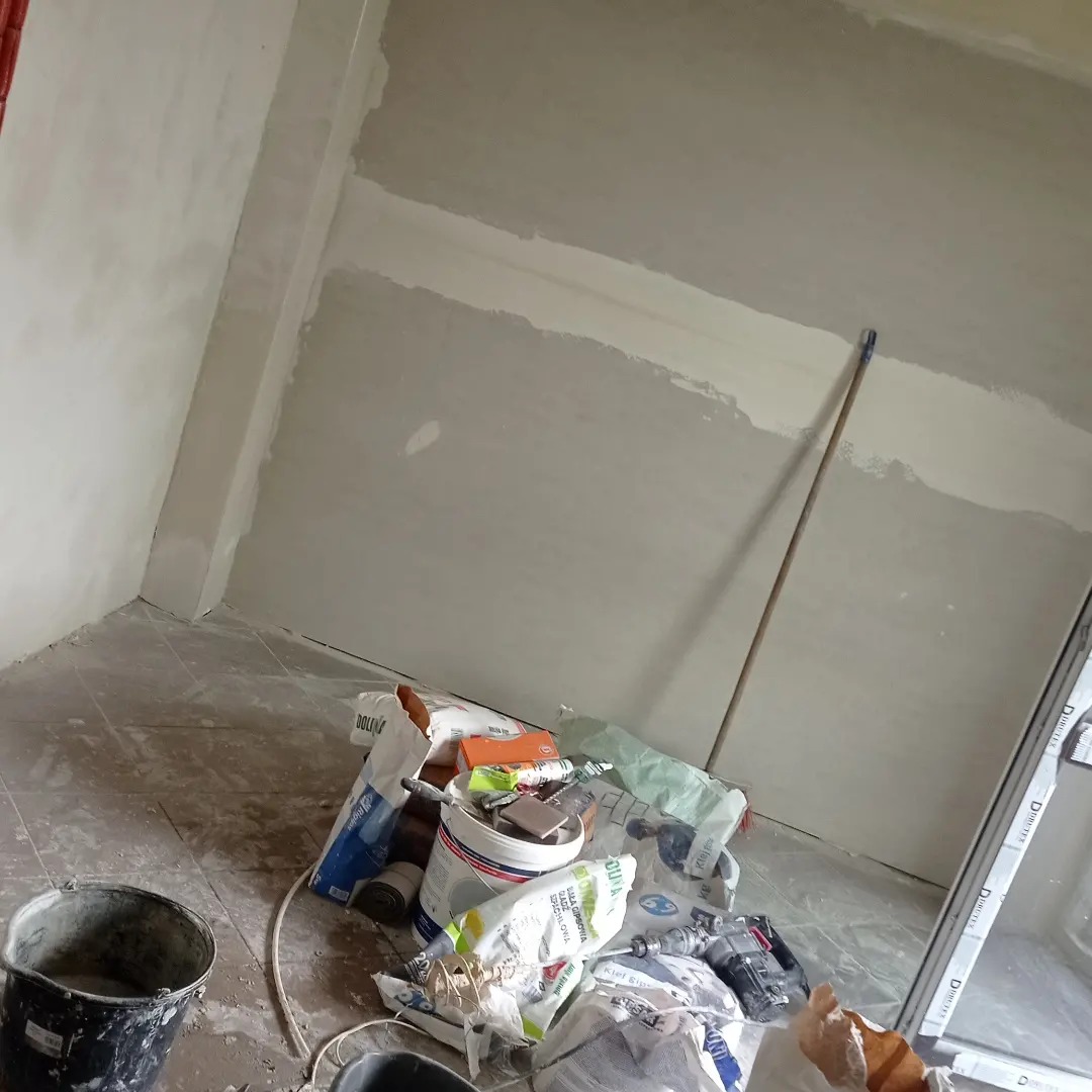 Przygotowanie ścian do malowania: częściowo zagruntowane ściany z widocznymi narzędziami i materiałami budowlanymi w nieuporządkowanym wnętrzu.
