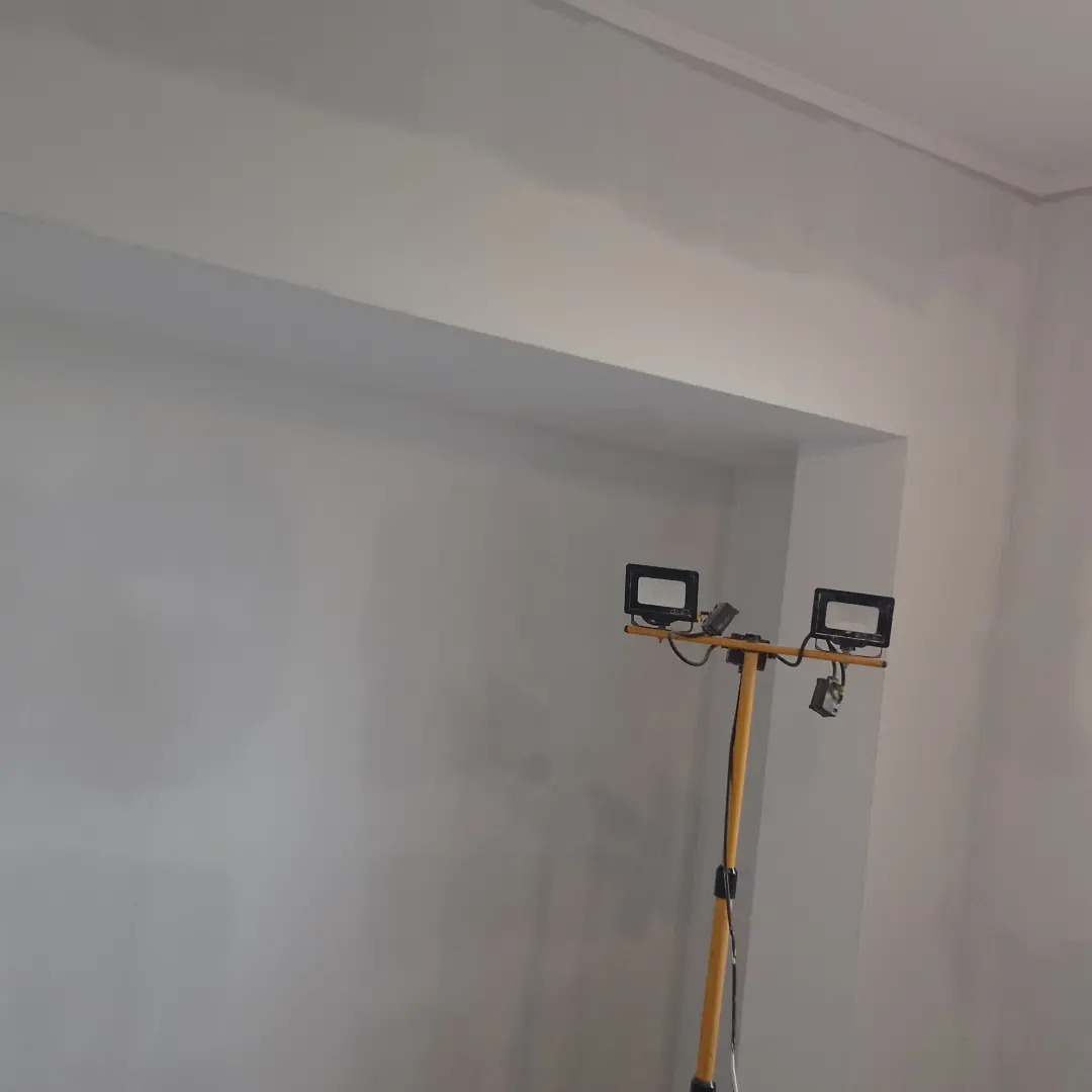 Świeżo pomalowana, szara ściana w rogu pokoju, oświetlona przez żółty statyw z dwoma halogenami.