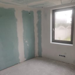 PWW - Puste pomieszczenie w trakcie prac wykończeniowych: ściany z zielonkawych płyt gipsowo-kartonowych z białymi spoinami, surowa podłoga, okno i wystające przewody elektryczne.