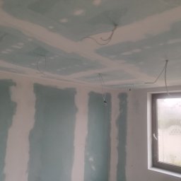 PWW - Pomieszczenie w trakcie prac wykończeniowych z zielonymi płytami na ścianach i suficie, widocznymi białymi spoinami oraz zwisającymi przewodami elektrycznymi. W prawym rogu okno z widokiem...