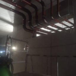 Hydrosfera instalacje - Czerwone rury instalacji grzewczej pod sufitem, widoczne wsporniki i obejmy, w tle surowy beton, na pierwszym planie butla z gazem i rusztowanie.