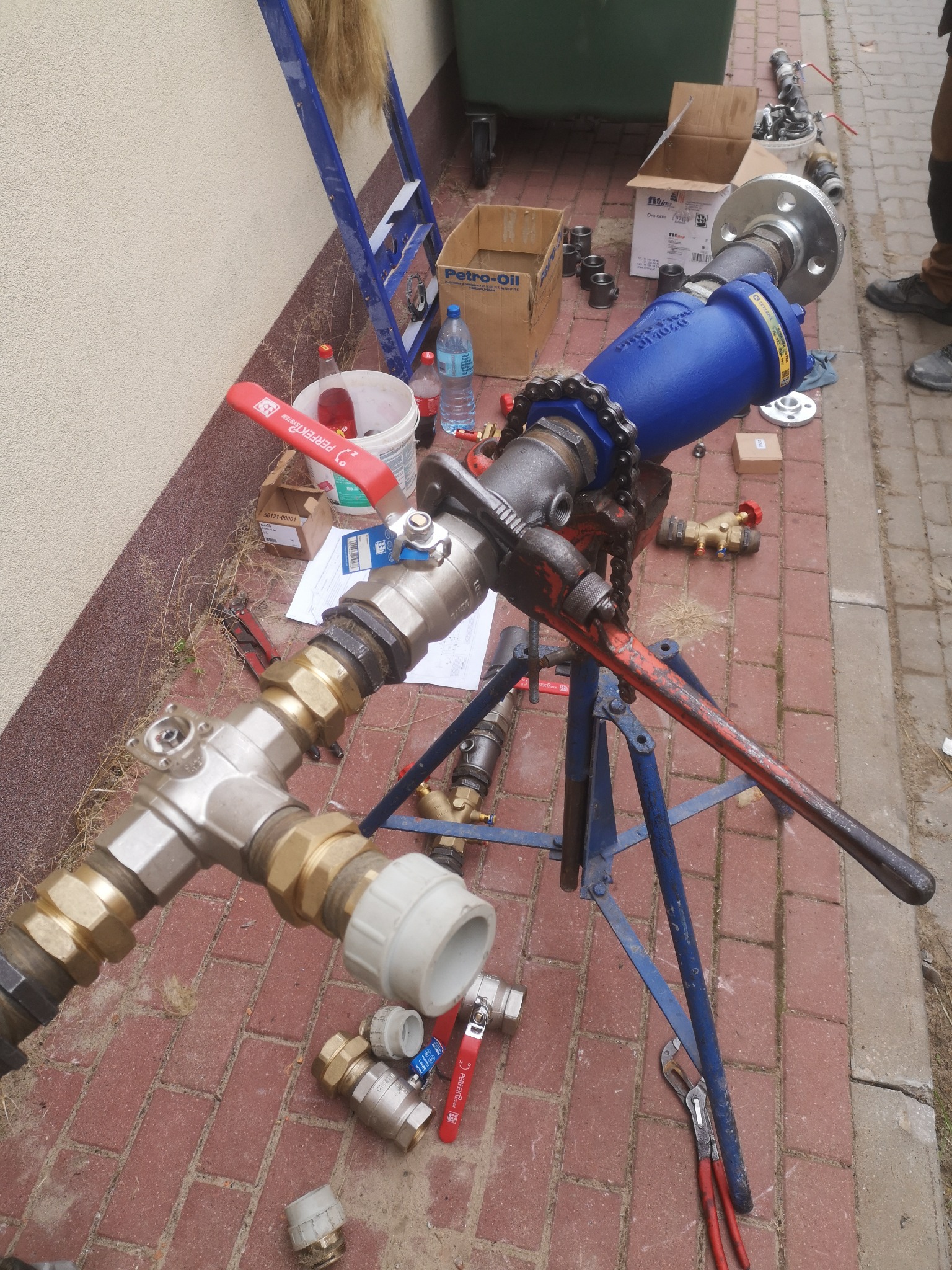 Elementy instalacji hydraulicznej na ceglanym podłożu, z widocznym niebieskim filtrem, rurami, zaworami, kluczem do rur i innymi narzędziami, sugerujące etap montażu lub przeróbki.