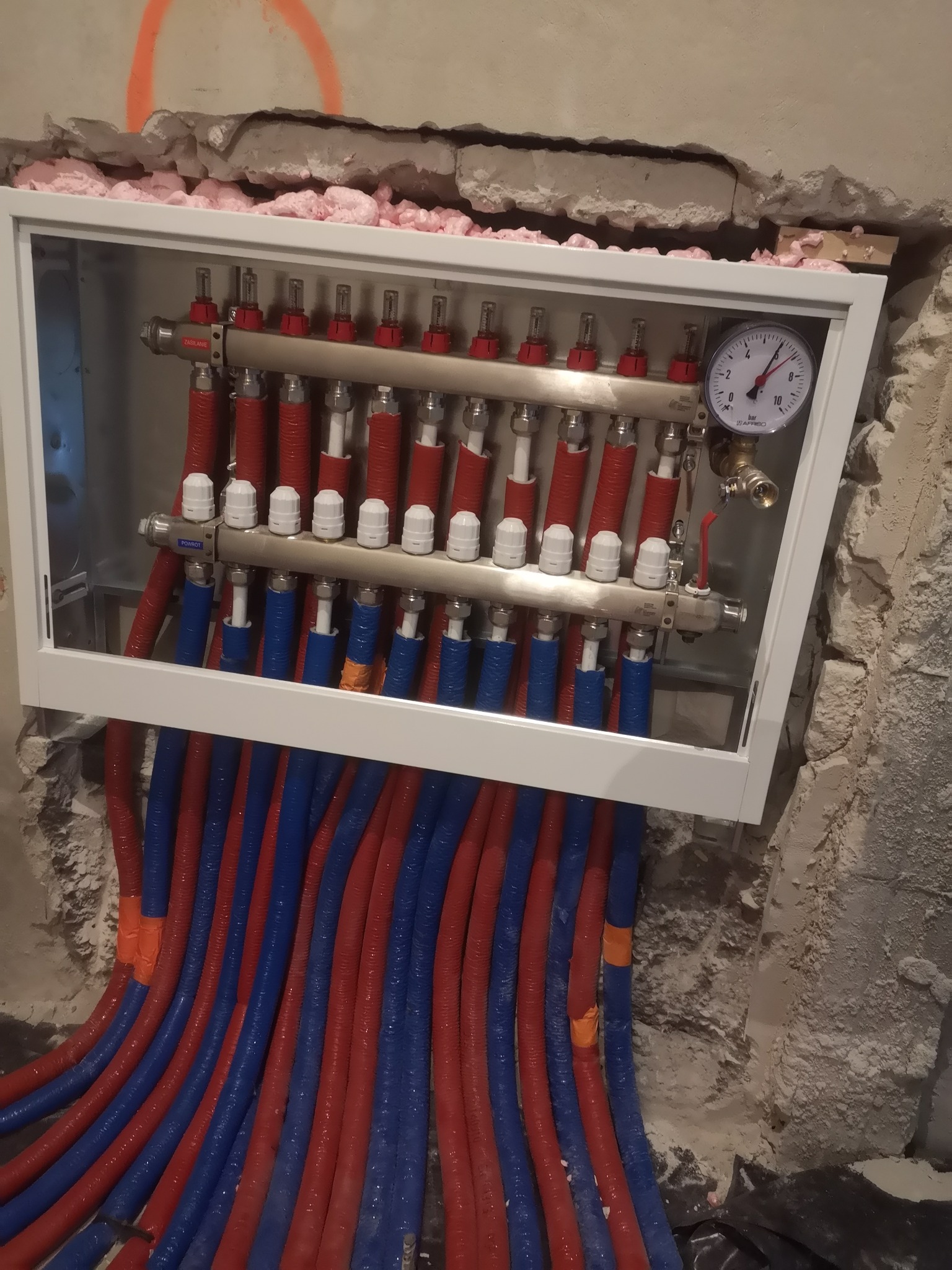 Rozdzielacz instalacji grzewczej z manometrem, zamontowany w ścianie, z widocznymi rurami w kolorach czerwonym i niebieskim oraz pianką uszczelniającą wokół ramy.