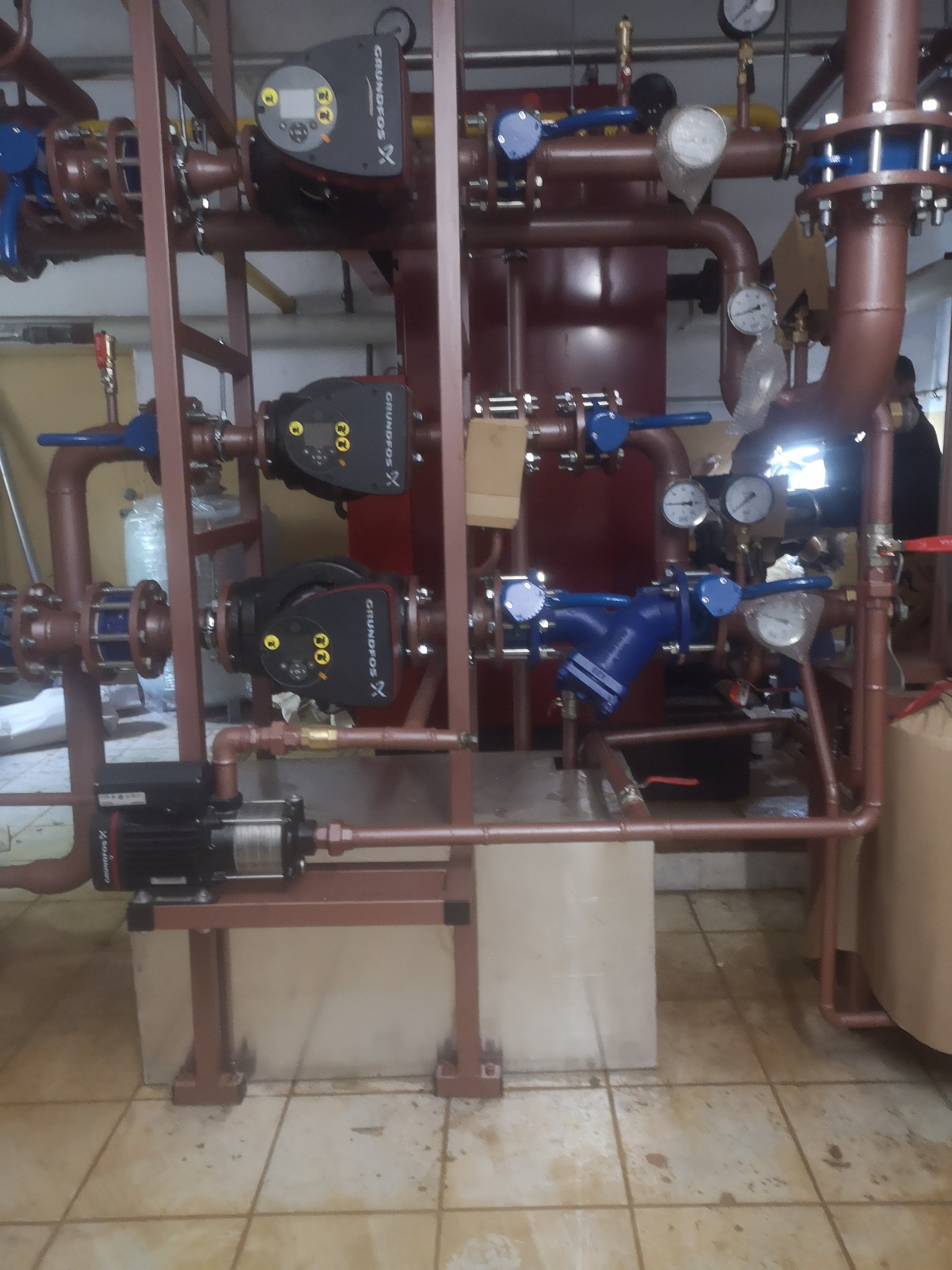 Rozbudowana instalacja grzewcza z kilkoma pompami obiegowymi Grundfos, zaworami, rurami w kolorze miedzianym i manometrami, zamontowana w pomieszczeniu technicznym.