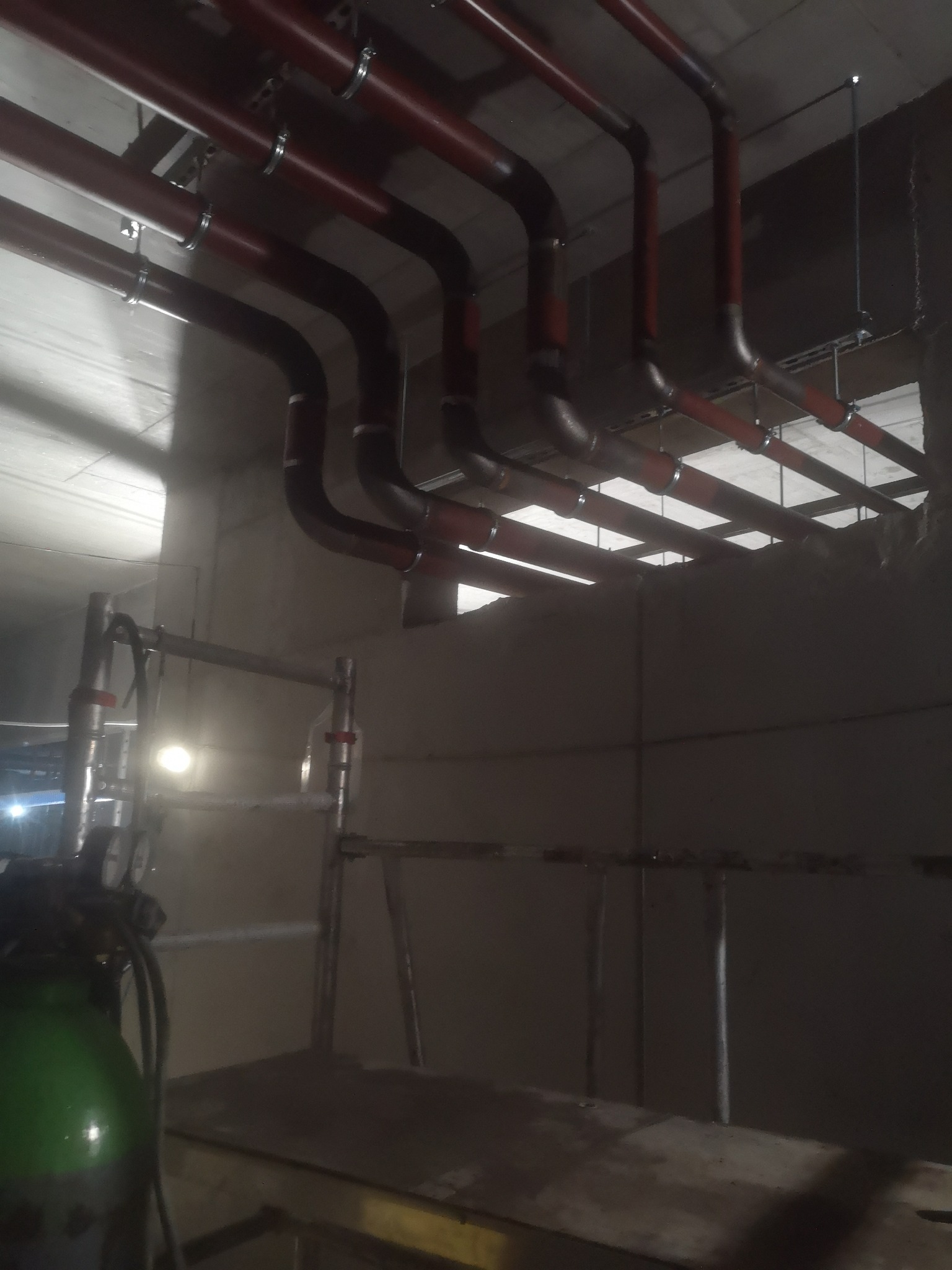 Czerwone rury instalacji grzewczej pod sufitem, widoczne wsporniki i obejmy, w tle surowy beton, na pierwszym planie butla z gazem i rusztowanie.