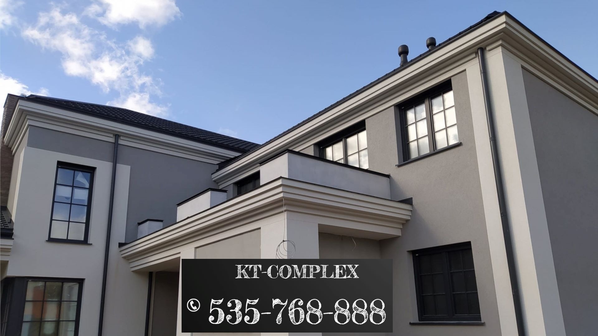 Dwukondygnacyjny budynek z szarą elewacją, czarną stolarką okienną i dachem, widoczny fragment nieba, na pierwszym planie czarne pole z białym napisem KT-COMPLEX i numerem telefonu.
