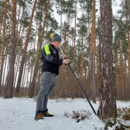 Pracownik w Tomaszowie Mazowieckim w zimowej scenerii, ustawia tyczkę geodezyjną w śniegu, na tle sosnowego lasu.