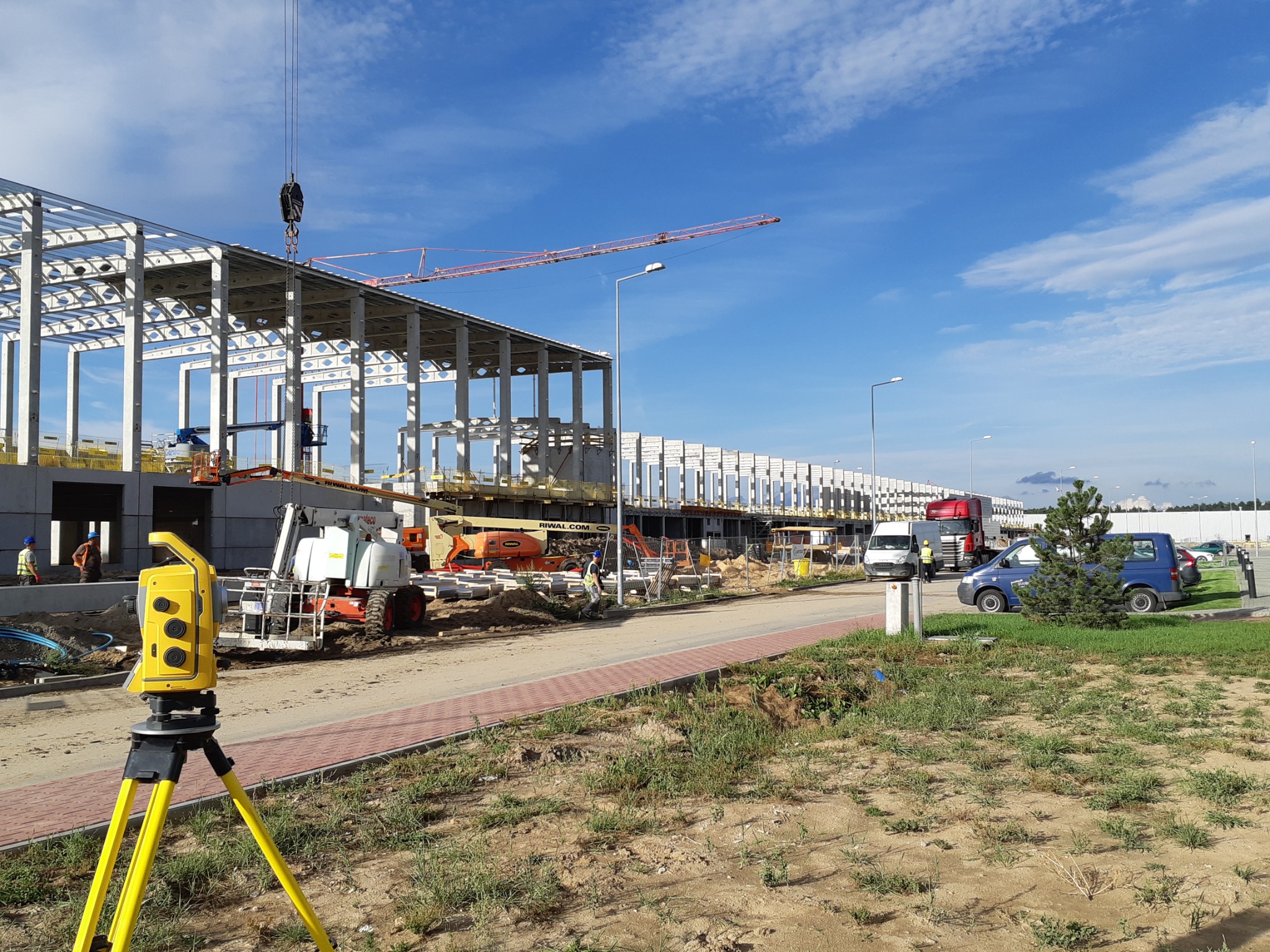 Żółty teodolit na trójnogu na pierwszym planie, w tle budowa hali magazynowej z konstrukcją stalową, żurawiem i podnośnikami koszowymi.