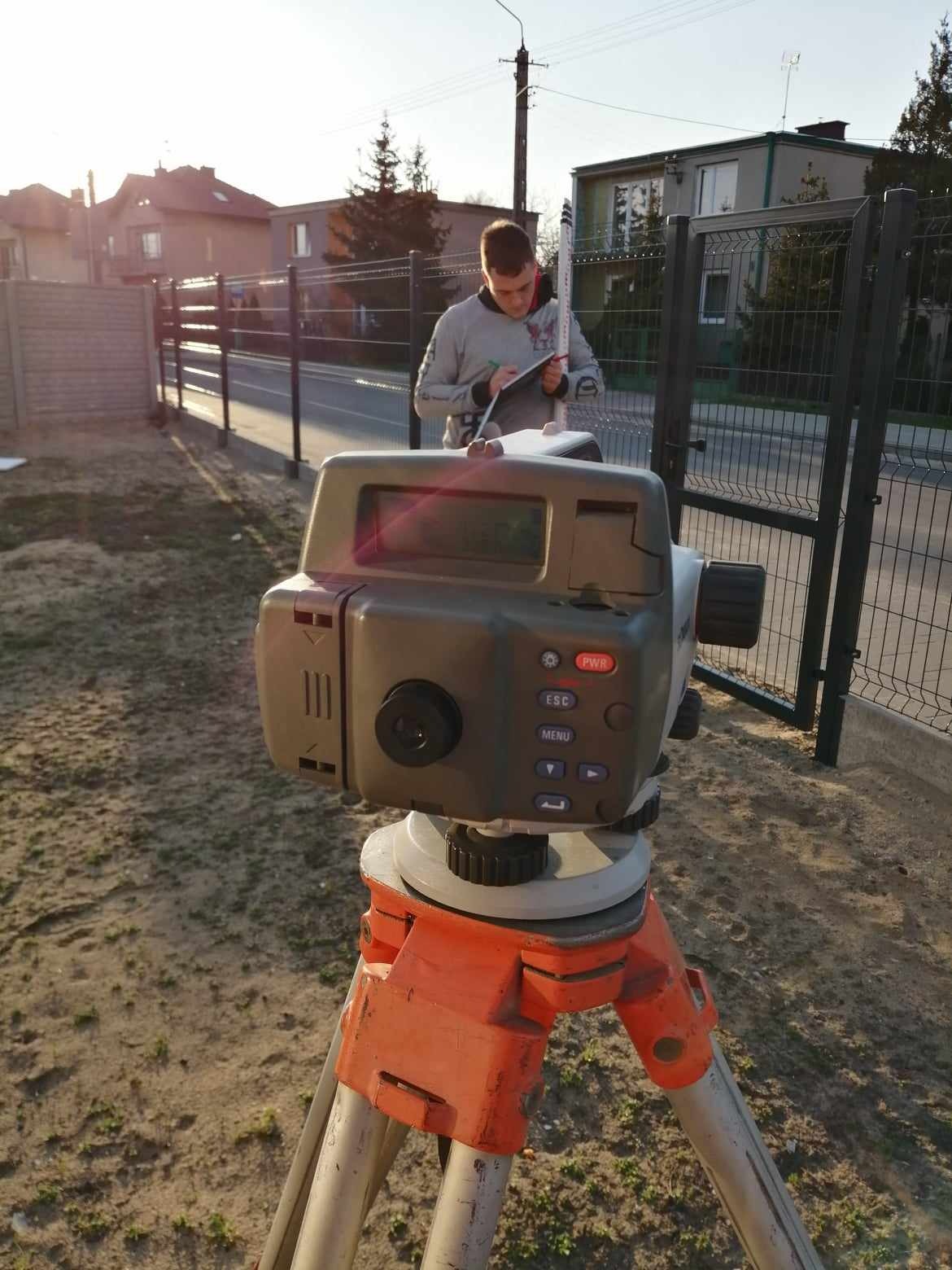 Geodeta przy pracy z niwelatorem elektronicznym na pomarańczowym statywie, notujący wyniki pomiarów na tle ogrodzenia i zabudowy jednorodzinnej.